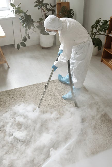 A close-up view of a domestic vacuum cleaner with a clear dust container filled with debris, positioned on a pink carpeted floor in a well-lit room. The vacuum's flexible hose, covered in a black and 