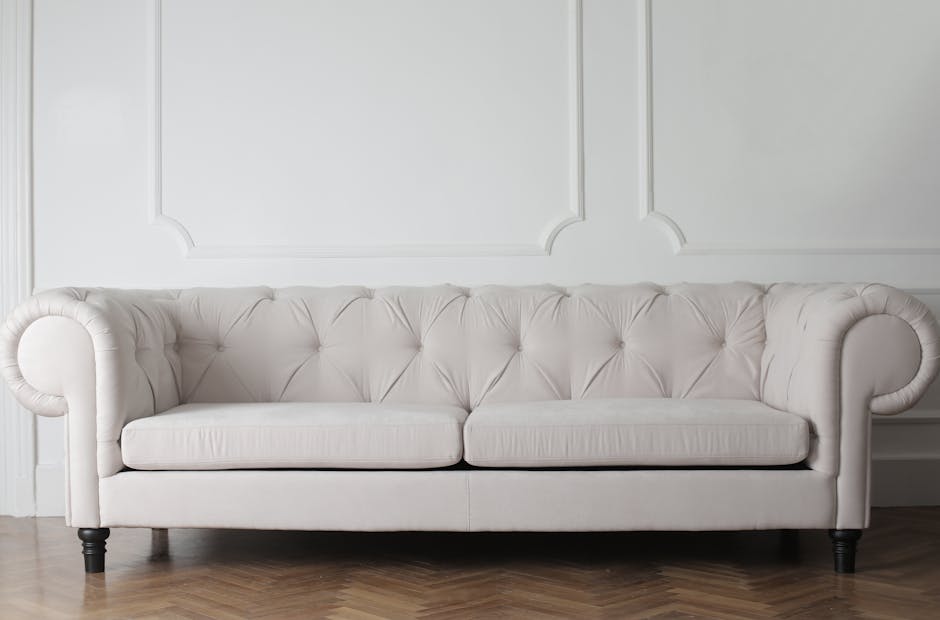 A modern living room featuring a light grey fabric sofa with matching cushions positioned against a minimalist wall with vertical paneling. To the left, there is a white kitchen counter with two black